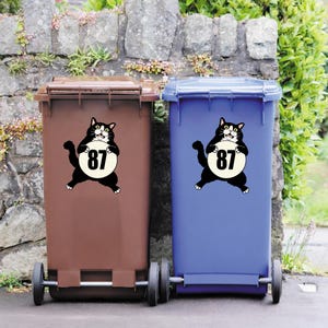 May include: Two wheeled trash bins, one brown and one blue, with a cartoon cat holding a sign with the number 87. The cat is black with white accents. The bins are outdoors, set against a stone wall and greenery.