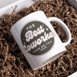 以下が含まれることがあります： 「The Best Coworker Gave Me This Mug」と書かれた茶色の円形デザインが施された白いセラミックマグカップです。