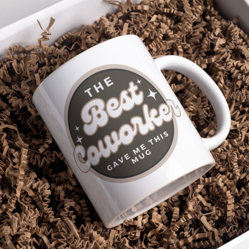 以下が含まれることがあります： 「The Best Coworker Gave Me This Mug」と書かれた茶色の円形デザインが施された白いセラミックマグカップです。