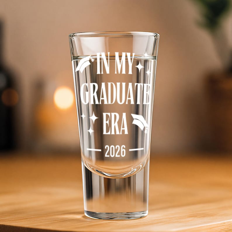 May include: Clear shot glass with the text "IN MY GRADUATE ERA 2026" in white. The glass has a graduation cap graphic. The shot glass is filled with a clear liquid and sits on a wooden surface.