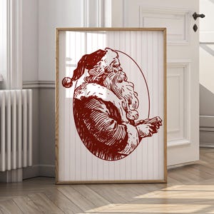 May include: A framed print featuring a vintage illustration of Santa Claus in a red and white color scheme. The artwork depicts Santa with a long white beard and a red hat, set against a striped background. The frame is made of light-colored wood.