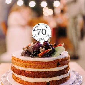 May include: White cake with white frosting and a round cake topper that says "Sue is 70" with a star and a banner.