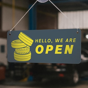 May include: A rectangular sign with the words "HELLO, WE ARE OPEN" in yellow, along with a stack of yellow tires. The sign is dark gray and hangs from a clear string, indicating a business is open.