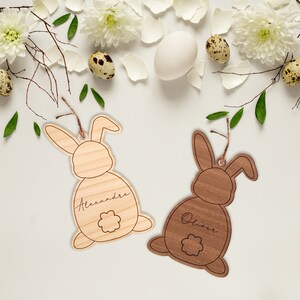 May include: Two wooden bunny shaped ornaments with the names "Alexandre" and "Oliver" engraved on them. The bunnies are brown and light brown and have a flower design on their backs.