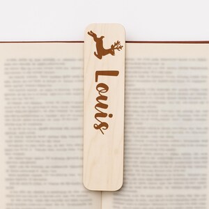 May include: A wooden bookmark with a brown reindeer illustration and the name "Louis" in a matching brown script. The bookmark is rectangular with rounded edges and is placed in an open book.