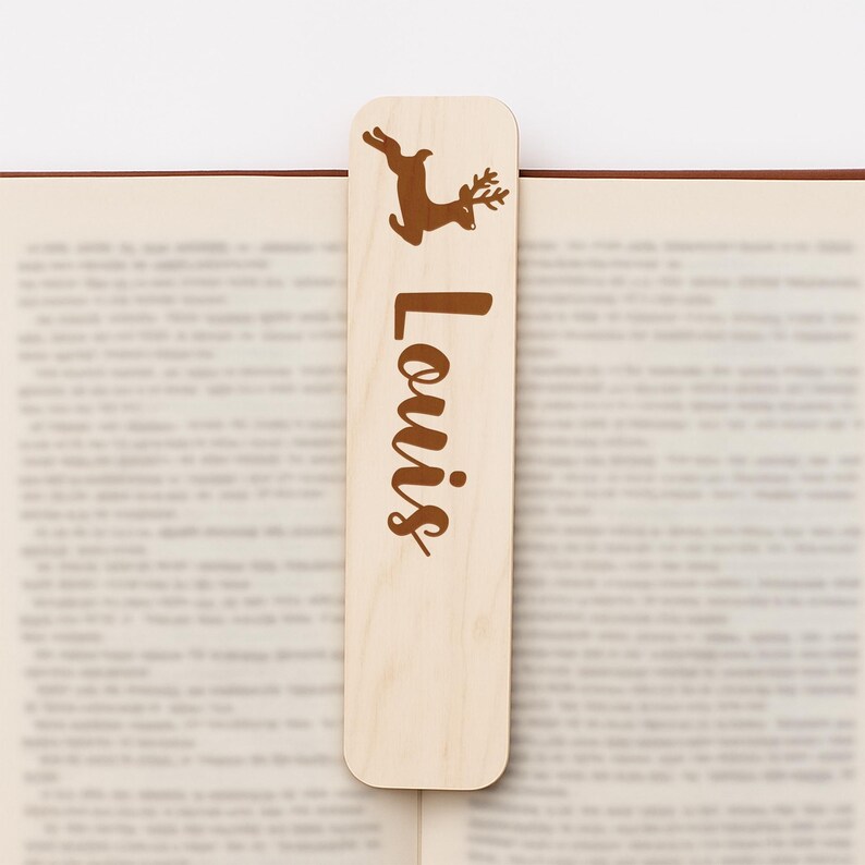 May include: A wooden bookmark with a brown reindeer illustration and the name "Louis" in a matching brown script. The bookmark is rectangular with rounded edges and is placed in an open book.