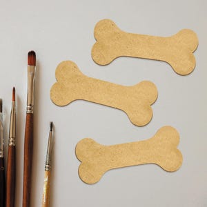 May include: Three brown cardboard dog bone shapes on a white surface. Five paintbrushes are arranged vertically on the left side of the image.