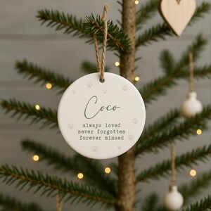 May include: A round, white ceramic Christmas ornament with the name "Coco" in script. The ornament has paw prints and the text "always loved, never forgotten, forever missed". It hangs from a twine loop.