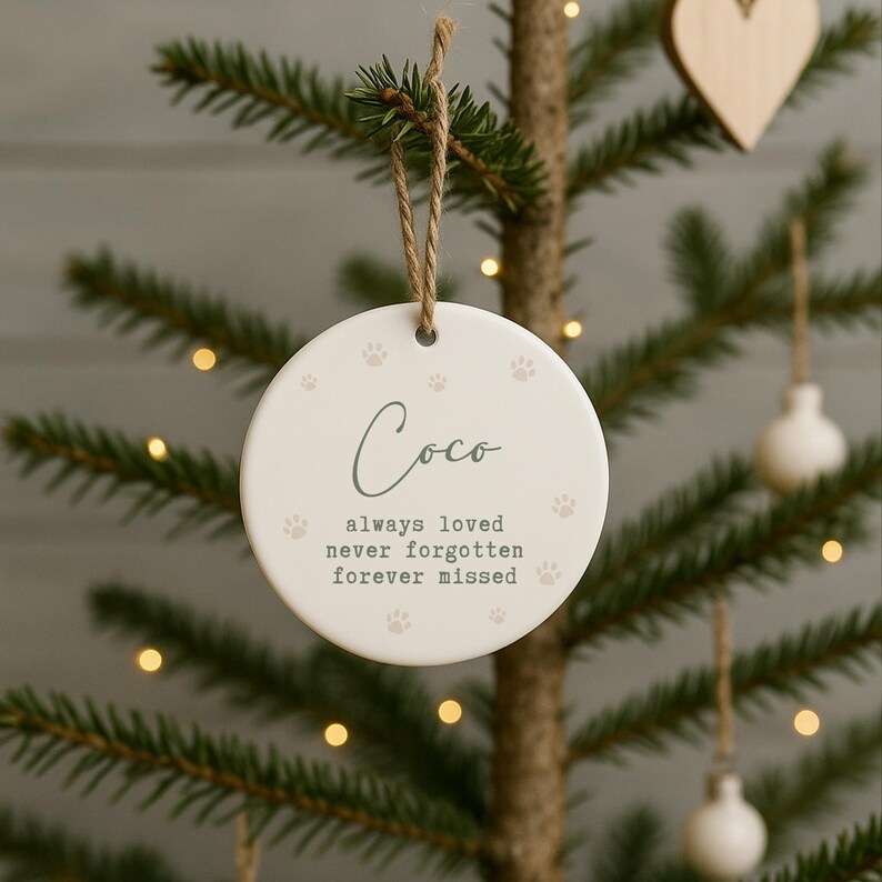 May include: A round, white ceramic Christmas ornament with the name "Coco" in script. The ornament has paw prints and the text "always loved, never forgotten, forever missed". It hangs from a twine loop.