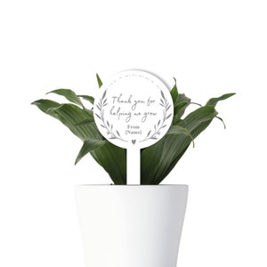 May include: White plant stake with a round, gray, floral wreath design. The stake says "Thank you for helping me grow. From [Name]"