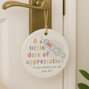 May include: A round, white ceramic door hanger with the text "A little dose of appreciation to say thanks for all you do!" in colorful letters. A syringe illustration is also featured.