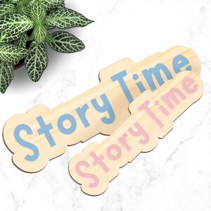 May include: Two wooden signs with the words "Story Time" in blue and pink lettering. The signs are light brown with a cloud-like shape. A green plant is visible in the upper left corner.
