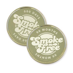 May include: Two gold-colored round tokens with a white circle in the center. The circle has the text "Smoke Free" in a retro font. One token has the text "000 Days" around the circle and the other has the text "00 Months" around the circle.