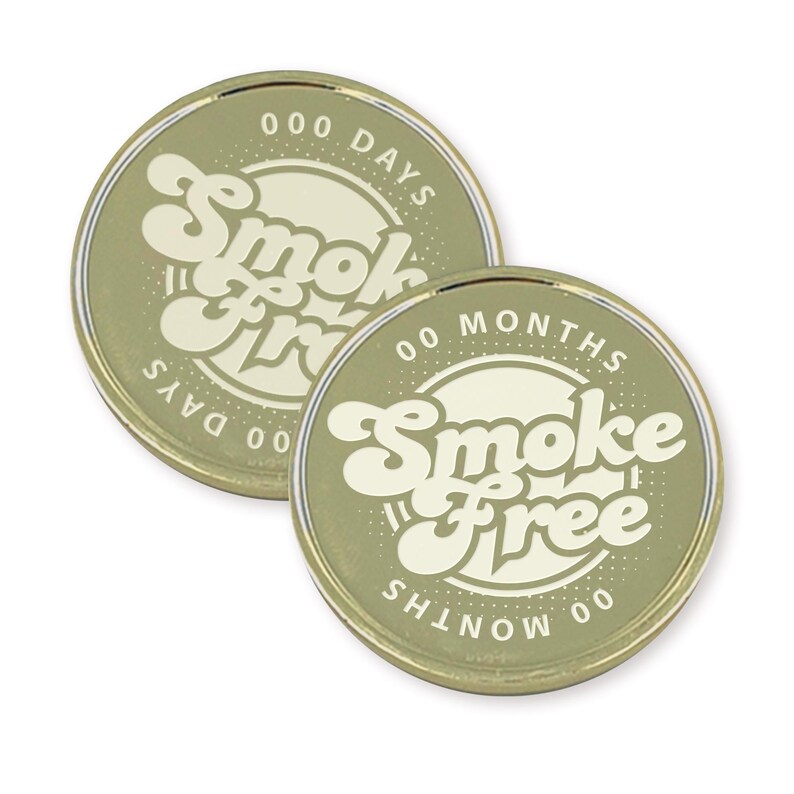 May include: Two gold-colored round tokens with a white circle in the center. The circle has the text "Smoke Free" in a retro font. One token has the text "000 Days" around the circle and the other has the text "00 Months" around the circle.