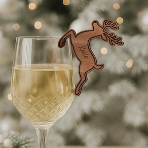 May include: A wooden reindeer wine charm with the name "Rosie" engraved on it, hanging on the rim of a wine glass filled with white wine. The charm is brown and has a detailed design. The background is blurred with soft, out-of-focus lights.