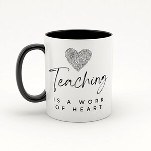Puede incluir: Taza de cer&aacute;mica blanca con asa e interior negros. La taza presenta el texto "Teaching is a work of heart" en escritura negra, junto con un dise&ntilde;o de coraz&oacute;n de huellas dactilares. Un regalo atento para profesores.