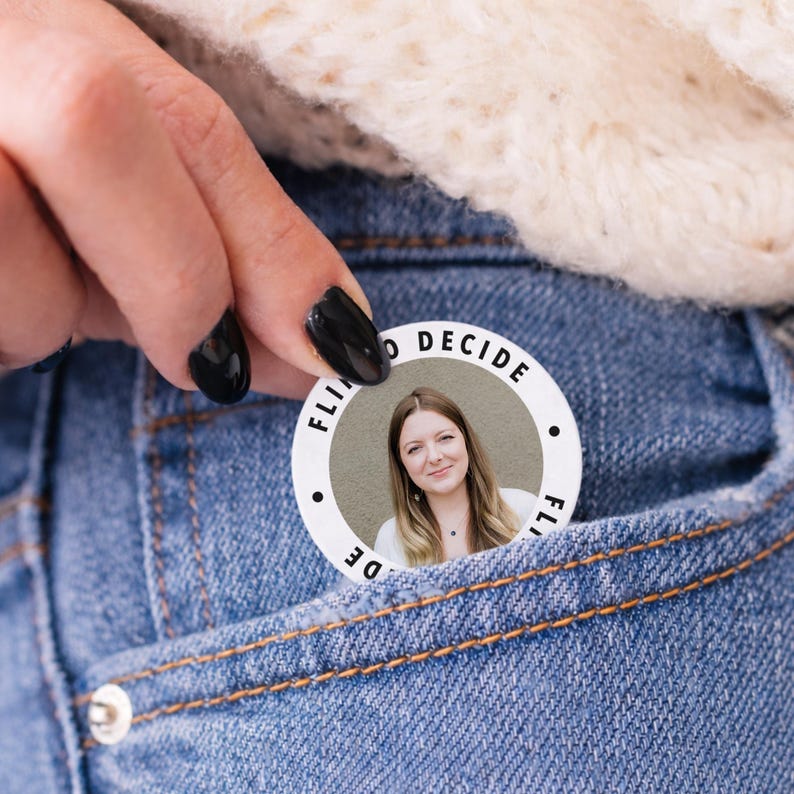 May include: A white button with the text "FLIP TO DECIDE" encircling a photo of a woman. The button is held in a denim pocket. The woman in the photo is smiling and wearing a white shirt.