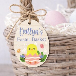 May include: Wooden Easter basket tag in an egg shape, with the text "Kaitlyn's Easter Basket" in blue script. The tag features a yellow chick hatching from a pink egg, surrounded by flowers and an Easter egg design. The tag is attached to a woven basket.