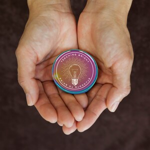 Puede incluir: Un pin redondo e iridiscente con el texto "Teaching Bright Students to Shine" que rodea un gr&aacute;fico de bombilla. El pin se sostiene en manos abiertas sobre un fondo marr&oacute;n.