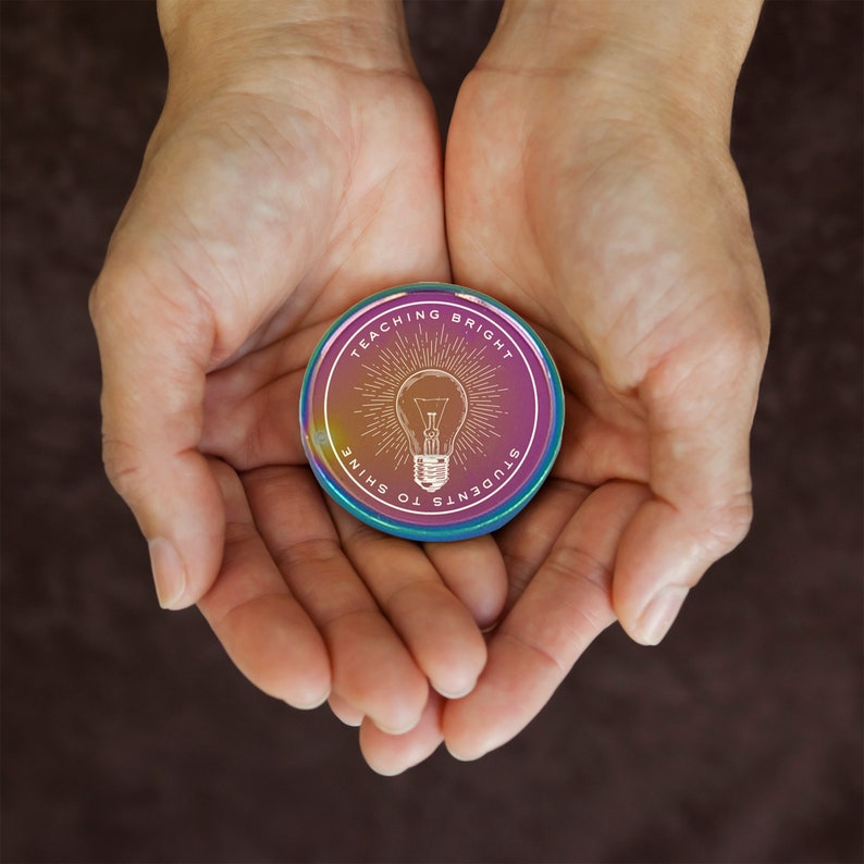 Puede incluir: Un pin redondo e iridiscente con el texto "Teaching Bright Students to Shine" que rodea un gr&aacute;fico de bombilla. El pin se sostiene en manos abiertas sobre un fondo marr&oacute;n.