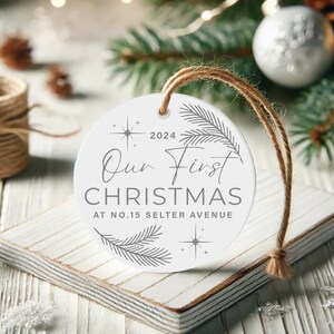 Op de afbeelding: Ronde witte kerstbal met een grijs ontwerp. De kerstbal heeft de tekst "Our First Christmas at No. 15 Selter Avenue" en het jaar "2024".