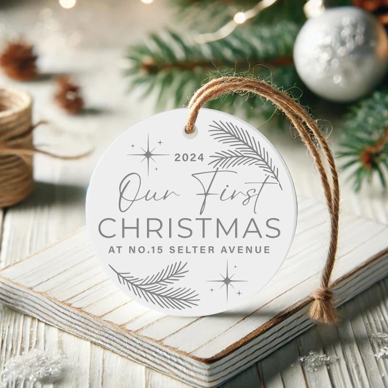 Op de afbeelding: Ronde witte kerstbal met een grijs ontwerp. De kerstbal heeft de tekst "Our First Christmas at No. 15 Selter Avenue" en het jaar "2024".