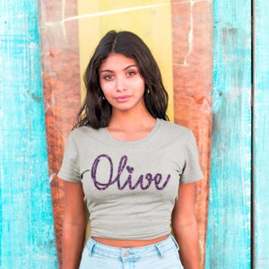 May include: A light gray short-sleeved t-shirt featuring the word Olive written in a dark purple glittery script font across the chest. The letter i in the word is topped with a small heart shape instead of a standard dot, centered on the shirt.