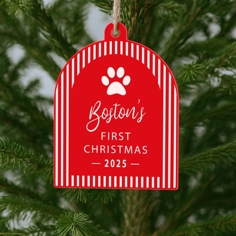 May include: Red Christmas ornament with white vertical stripes and a paw print. The ornament reads "Boston's First Christmas - 2025" in white script. The ornament is hanging from a natural twine string.