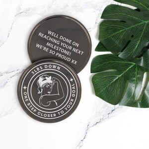 Puede incluir: Dos posavasos redondos negros con texto blanco. El posavasos superior dice "Well done on reaching your next milestone! We're so proud xx". El posavasos inferior tiene una ilustraci&oacute;n en blanco y negro de una mujer flexionando el b&iacute;ceps con el texto "5 lbs down one step closer to your goal".