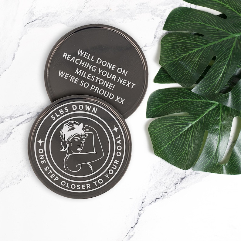 Puede incluir: Dos posavasos redondos negros con texto blanco. El posavasos superior dice "Well done on reaching your next milestone! We're so proud xx". El posavasos inferior tiene una ilustraci&oacute;n en blanco y negro de una mujer flexionando el b&iacute;ceps con el texto "5 lbs down one step closer to your goal".