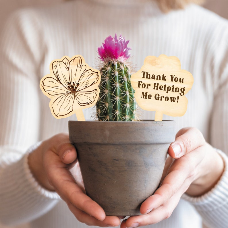May include: A small cactus in a grey pot with a pink flower and two wooden signs. One sign is a flower outline, and the other says "Thank You For Helping Me Grow!" The pot is held in both hands.