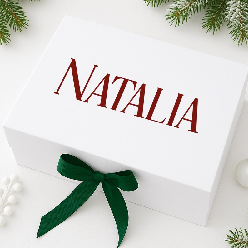 May include: A white gift box with the name "NATALIA" printed in a deep red color on the lid. A dark green ribbon is tied in a bow on the front of the box. The box is decorated with Christmas greenery.