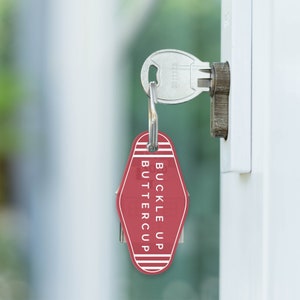 Puede incluir: Una etiqueta de llave de motel roja con texto blanco que dice "Buckle Up Buttercup". La etiqueta de llave est&aacute; colgada de una llave plateada en una cerradura de puerta.