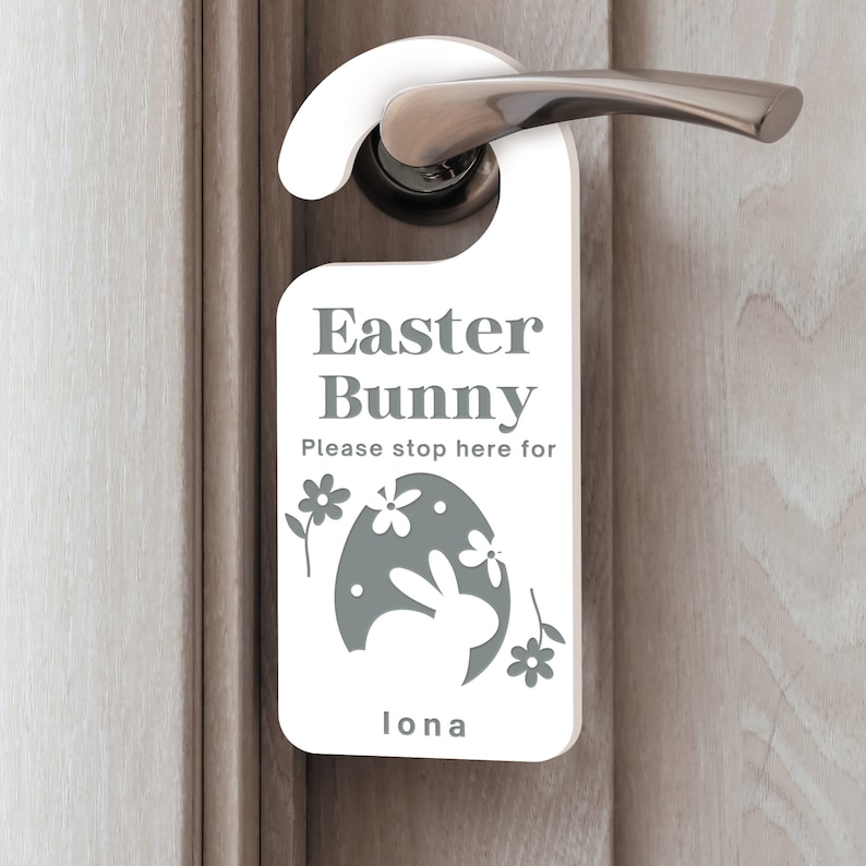 May include: White door hanger with the text "Easter Bunny" and "Please stop here for" with a gray silhouette of a bunny rabbit inside an egg. The text "Iona" is at the bottom of the hanger.