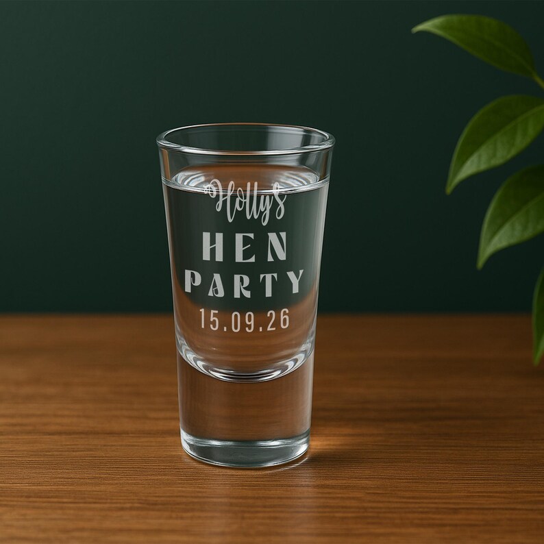 Op de afbeelding: Een helder shotglaasje gevuld met vloeistof, met de tekst "Holly's HEN PARTY 15.09.26". Het glas staat op een houten oppervlak tegen een donkergroene achtergrond, met een plant rechts. Perfect voor een vrijgezellenfeest.