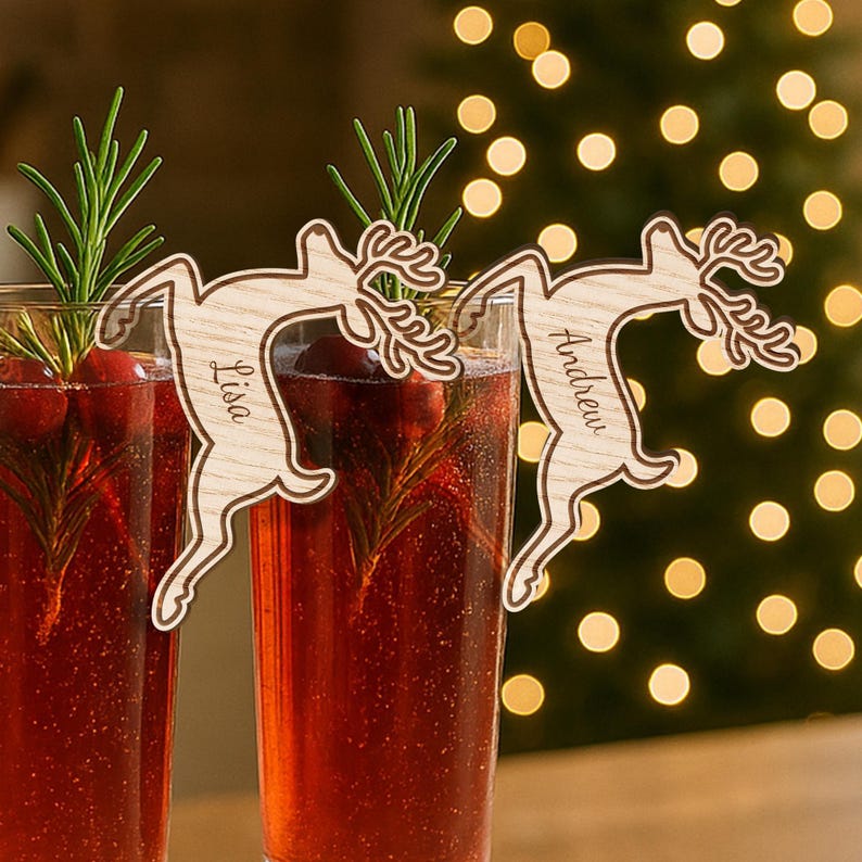 May include: Two glasses of a red beverage, garnished with rosemary and cranberries, are adorned with wooden reindeer drink stirrers. One stirrer has the name "Lisa" and the other "Andrew" engraved on it. The background features blurred Christmas lights.