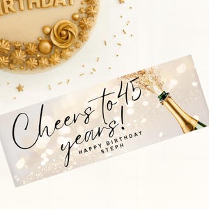 以下が含まれることがあります： 「Cheers to 45 years! Happy Birthday Steph」の文字と、金色のスプレーが付いたシャンパンボトルのバナー。バナーは白と金のボケの背景です。金色の装飾が施されたケーキが左上にあります。