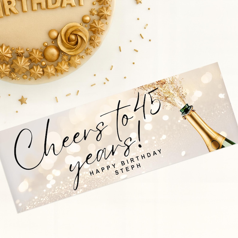 以下が含まれることがあります： 「Cheers to 45 years! Happy Birthday Steph」の文字と、金色のスプレーが付いたシャンパンボトルのバナー。バナーは白と金のボケの背景です。金色の装飾が施されたケーキが左上にあります。