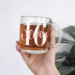 May include: A clear glass mug filled with a brown liquid, featuring the number "16" in white and the name "Olivia" in pink script. The mug is held by a person, with a green plant visible in the background.