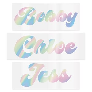 Puede incluir: Tres pegatinas rectangulares con los nombres "Bobby", "Chloe" y "Jess". Cada nombre est&aacute; escrito en una fuente cursiva, estilo burbuja, con un efecto hologr&aacute;fico de arco&iacute;ris. Las pegatinas est&aacute;n sobre un fondo blanco.