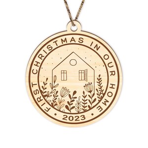 May include: A round wooden ornament with the text "First Christmas in Our Home 2023" and a house with a window and a door, surrounded by stylized plants.