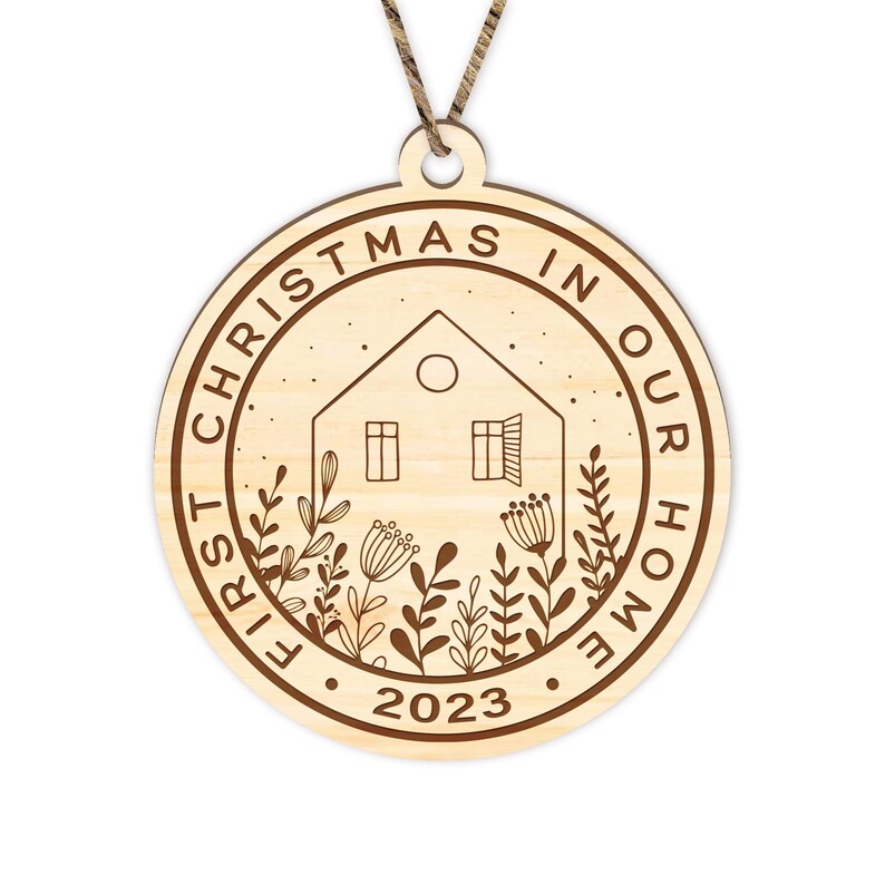 May include: A round wooden ornament with the text "First Christmas in Our Home 2023" and a house with a window and a door, surrounded by stylized plants.