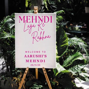 以下が含まれることがあります： ピンク色の筆記体で「MEHNDI Laga Ke Rakhna」と書かれた白い看板が木製のイーゼルに立てられています。「WELCOME TO AARUSHI'S MEHNDI 25/11/25」と書かれています。看板は緑豊かな植物に囲まれています。