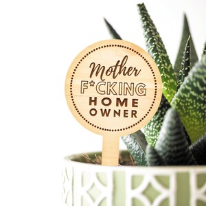 Puede incluir: Una estaca de planta de madera con un letrero redondo marr&oacute;n que dice "Mother F*CKING HOME OWNER". La estaca est&aacute; en una planta verde en maceta.