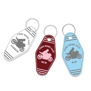 Op de afbeelding: Drie sleutelhangers met een motorfiets thema in wit, bordeauxrood en lichtblauw. Elke sleutelhanger heeft een motorfietsafbeelding en de tekst "COME HOME SAFE". Er staat "ANDREW", "MUM" en "DAD" op.