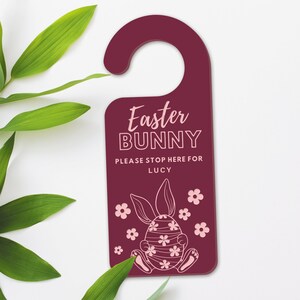 May include: Burgundy door hanger with the text "Easter BUNNY PLEASE STOP HERE FOR LUCY". The design includes a bunny illustration with floral accents. The hanger is surrounded by green leaves.