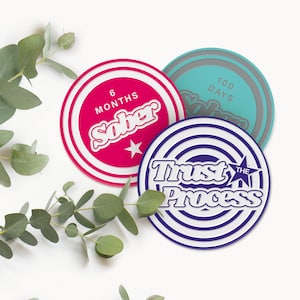 May include: Three round coasters with motivational messages. The first coaster is pink with white text that reads "6 Months Sober". The second coaster is blue with white text that reads "100 Days Sober". The third coaster is purple with white text that reads "Trust The Process".