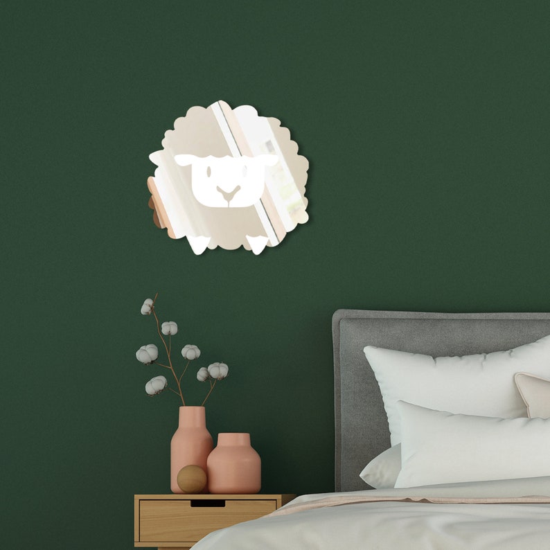 May include: A sheep-shaped mirror with a white face and a fluffy, beige body is mounted on a dark green wall. The mirror is a decorative accent, suitable for a nursery or children's room. A nightstand with pink vases and a bed are in the background.