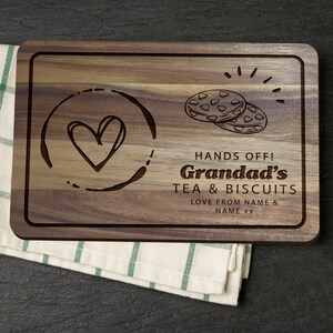 May include: A wooden cutting board with a heart and two biscuits engraved on it. The text reads "Hands Off! Grandad's Tea & Biscuits Love from NAME & NAME XX".