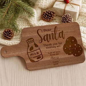 May include: A wooden cutting board with a handle, engraved with "Dear Santa" and a milk bottle and cookies illustration. The text reads "Thank you for visiting us! Lots of love, from the Morris family xoxo."
